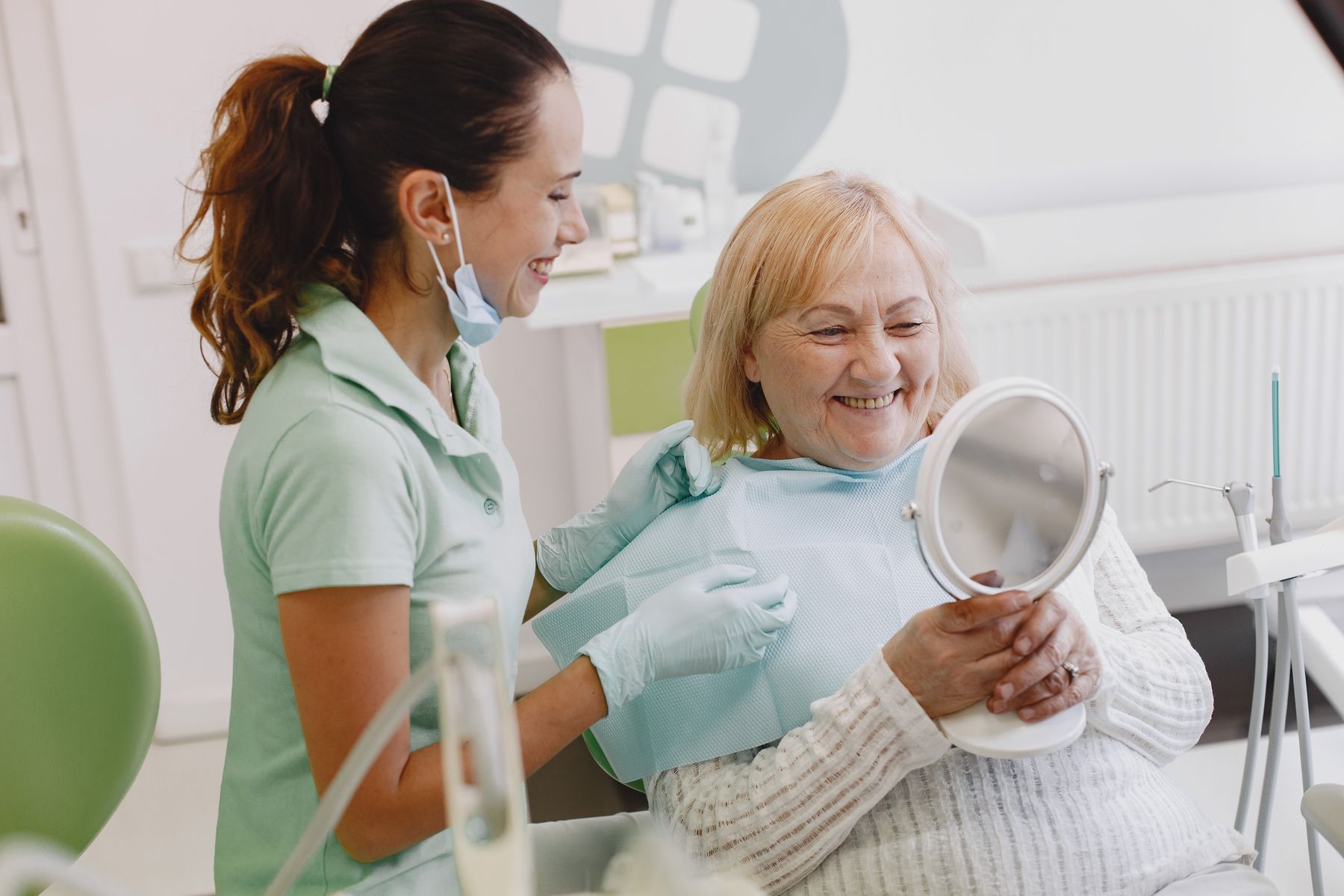 patiente aux cheveux blonds souriant dans un fauteuil de dentiste, miroir à la main, à côté de la praticienne